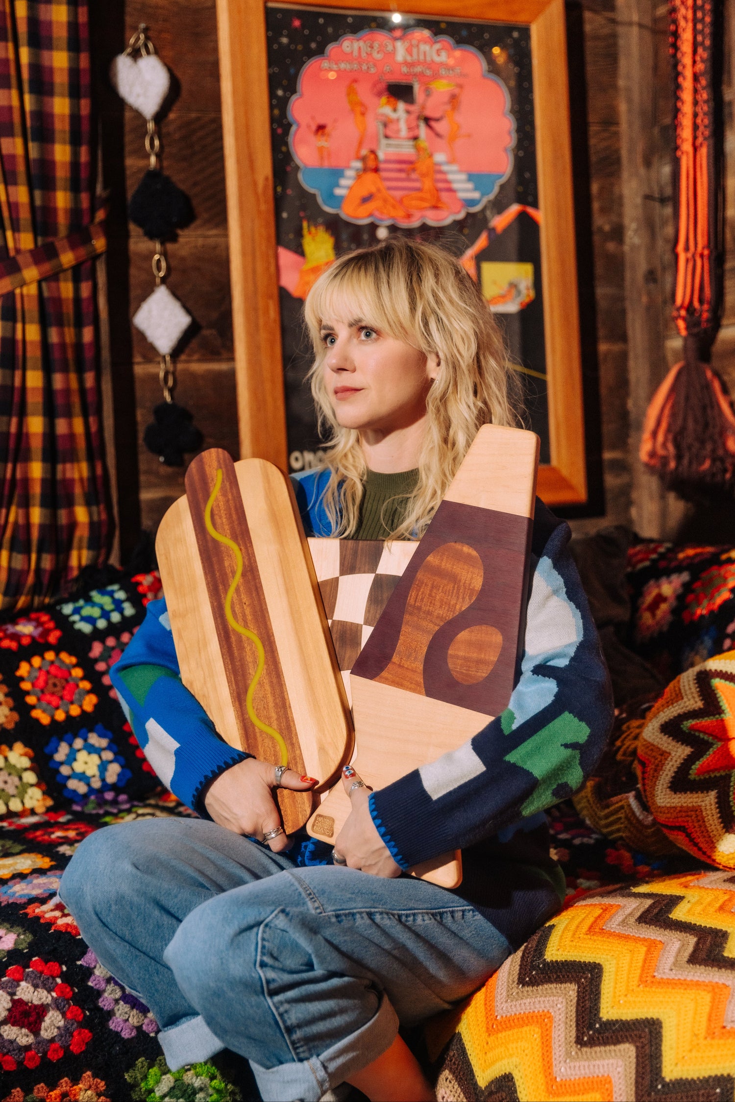 A woman sits on a colorful crocheted sofa holding three wooden cutting boards with different patterns, in a cozy, rustic room with a framed art print on the wall.