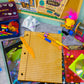 A styled flat-lay of school supplies and party accessories on a gray surface, including a wooden lined notebook, colorful pencils, a purple school picture box, a yellow 'Handy Dandy Dance' card, a toy, and other party and stationery items.