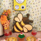 A styled tabletop scene with a yellow wall clock and a wooden bee-shaped object against floral wallpaper, with bowls of food, napkins, and drink glasses on a pink table.