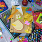 A styled flat-lay of assorted colorful stationery and school supplies on a gray textured surface, including notebooks, pens, a ruler, sticky notes, a CD case, and small decorative items.
