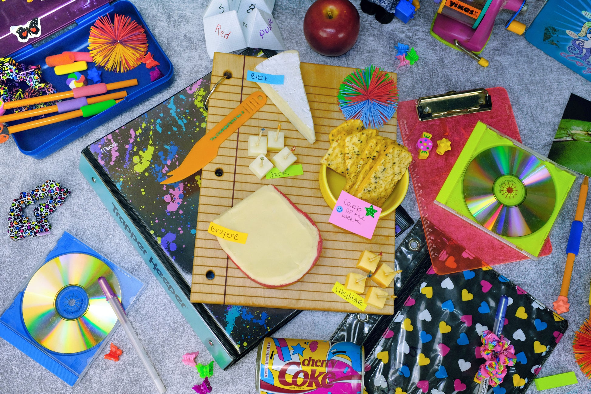 A styled flat-lay of assorted colorful stationery and school supplies on a gray textured surface, including notebooks, pens, a ruler, sticky notes, a CD case, and small decorative items.