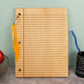 A wooden desk organizer with a lined notebook design, featuring a yellow plastic clip holding a pen, set on a gray surface against a mint green wall with decorative props.